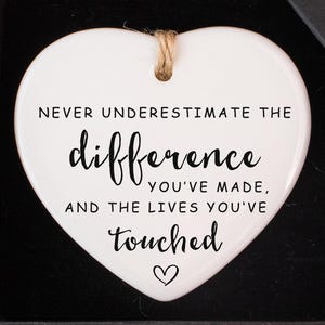 May include: A white heart-shaped ceramic ornament with the text "Never underestimate the difference you've made, and the lives you've touched" in black. The ornament is in a black gift box with a small blue flower decoration.