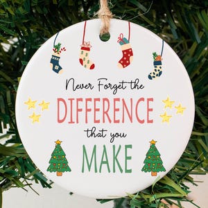 May include: A white, round Christmas ornament with the text "Never Forget the DIFFERENCE that you MAKE." The ornament features illustrations of stockings, stars, and Christmas trees. It hangs from a natural twine string.