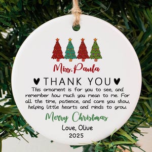 May include: A white ceramic Christmas ornament with a string for hanging. The ornament features four Christmas trees in red and green, the text "Miss Paula THANK YOU", and the words "Merry Christmas, Love, Olive 2025".