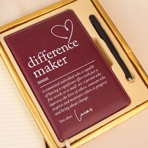 May include: A burgundy leather-bound journal with the words "difference maker" and a definition printed on the cover. The journal is in a gold box with a black pen. An open notebook and a vase of flowers are also in the image.