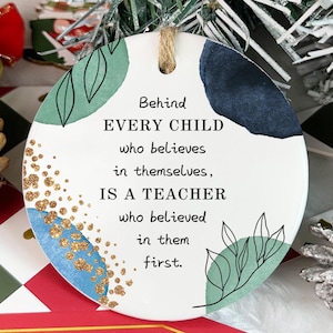 May include: White round ornament with blue, green, and gold abstract designs. The ornament has the text "Behind EVERY CHILD who believes in themselves, IS A TEACHER who believed in them first."