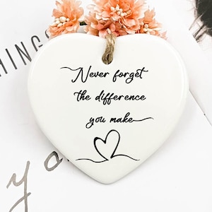 Op de afbeelding: Witte, hartvormige hanger van keramiek met de tekst "Vergeet nooit het verschil dat je maakt" en een klein hartontwerp. De hanger is versierd met roze bloemen.