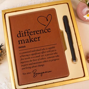 May include: A brown leather journal and black pen set in a gold box. The journal features the words "difference maker" and a definition. A heart graphic is above the text. The set is presented with flowers and a perfume bottle.