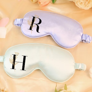 May include: Two satin sleep masks, one lavender and one mint green, are displayed. Each mask features a large initial, "R" and "H" respectively, in black with a floral design. The masks are surrounded by flowers and petals, suggesting a spa or relaxation theme.
