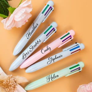 May include: Five multi-color pens in pastel shades of blue, pink, and green, each with a name printed on the barrel. The pens have a white top with colored buttons and are arranged on a peach-colored surface. A pink flower and greenery are in the background.