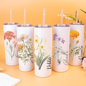 Op de afbeelding: Vijf witte roestvrijstalen bekers met bloemenontwerpen en namen. Elke beker heeft een andere bloemenillustratie, waaronder een anjer, een madeliefje, een narcis, een paarse bloem en een gele bloem. Elke beker heeft een doorzichtige rietje.