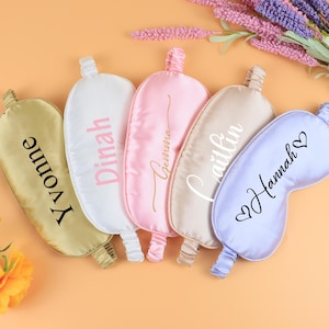 Könnte beinhalten: Fünf Schlafmasken aus Satin in Gold, Weiß, Rosa, Beige und Lavendel. Auf jeder Maske steht ein Name: Yvonne, Dinah, Gemma, Caitlin und Hannah. Die Masken sind auf einer orangefarbenen Oberfläche mit Blumen im Hintergrund angeordnet.