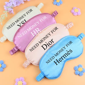 May include: Four satin sleep masks in cream, pink, peach, and blue, each with the text "NEED MONEY FOR" and a brand name. Decorative flowers and a sprig of purple flowers are scattered around the masks.