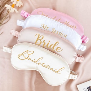 May include: Set of five satin sleep masks with gold lettering. The masks are in shades of pink and white. The text on the masks reads "Maid of Honor", "Mr. Jessica", "Bride", and "Bridesmaid".