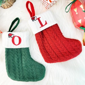 May include: Two festive Christmas stockings, one red and one green, with white knitted cuffs. The red stocking features a red embroidered "L", and the green stocking has a red "O". Both have a Santa hat detail and a hanging loop.