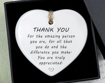 May include: A white ceramic heart-shaped ornament with a black string attached. The ornament has the text "Thank You" and a message of appreciation for an amazing person. The ornament is in a black gift box with a brown "Hand Made" tag.