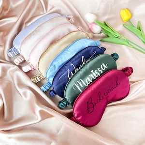 May include: A set of seven silk sleep masks in various colors, each with a personalized name or title embroidered in white. The masks are arranged in a stack, with the top mask reading "Bridesmaid".