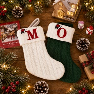May include: Two cable-knit Christmas stockings, one cream and one dark green, each featuring a white cuff with a red embroidered letter and a small red Santa hat icon. The cream stocking displays the letter M, and the green stocking displays the letter O.