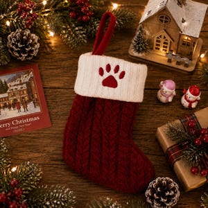 Peut inclure: Une chaussette de Noël en tricot torsadé rouge foncé avec un revers côtelé blanc orné d'une empreinte de patte brodée en rouge. La chaussette comporte une boucle en tissu rouge assortie sur le dessus pour la suspendre, avec un motif texturé.