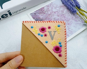 May include: A triangular felt corner bookmark in beige featuring red stitched edges. The surface is embroidered with a gray letter V, surrounded by small pink floral motifs and yellow and blue decorative stitching accents.