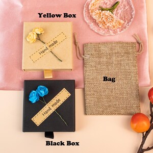 K&ouml;nnte beinhalten: Eine quadratische beigefarbene Schachtel mit zwei gelben Papierblumen und einem Etikett mit der Aufschrift Hand made, eine schwarze quadratische Schachtel mit zwei blauen Papierblumen und einem Etikett mit der Aufschrift Hand made sowie ein rechteckiger Jutebeutel mit Kordelzug.
