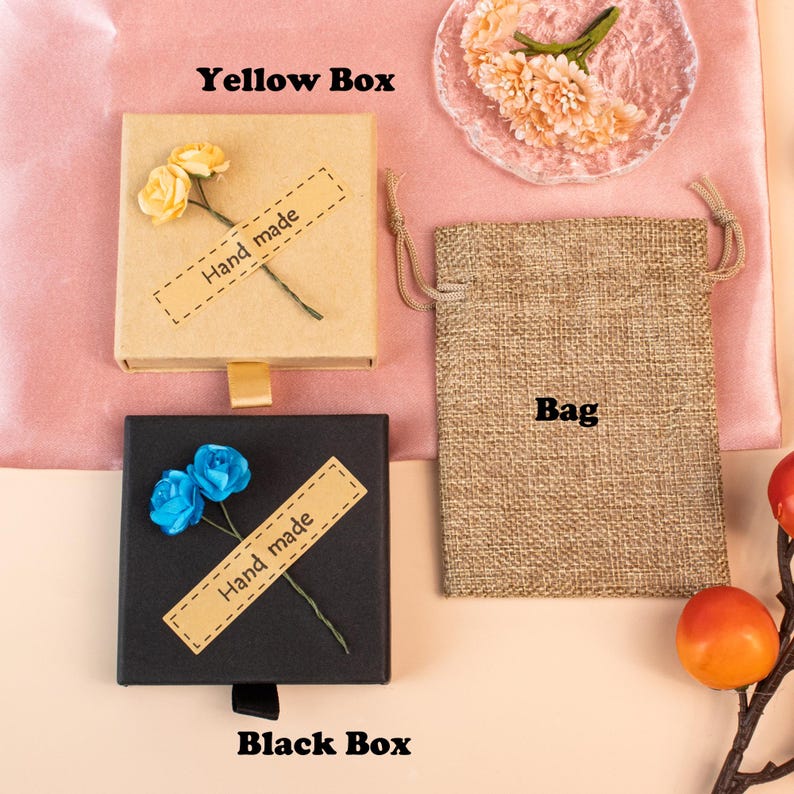 K&ouml;nnte beinhalten: Eine quadratische beigefarbene Schachtel mit zwei gelben Papierblumen und einem Etikett mit der Aufschrift Hand made, eine schwarze quadratische Schachtel mit zwei blauen Papierblumen und einem Etikett mit der Aufschrift Hand made sowie ein rechteckiger Jutebeutel mit Kordelzug.