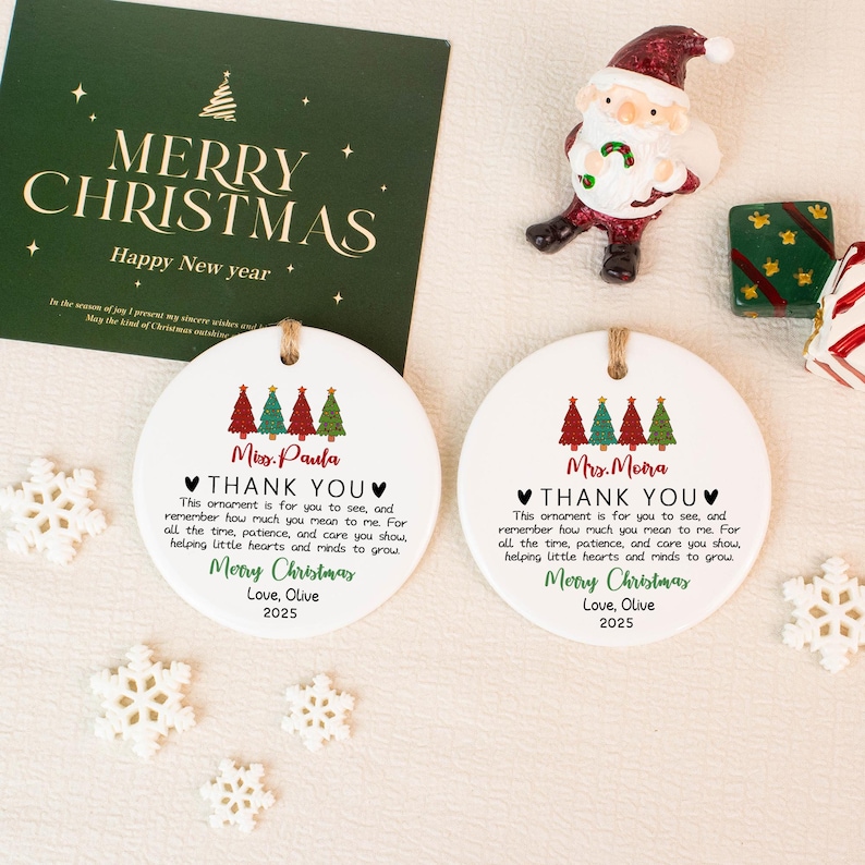 May include: Two white ceramic Christmas ornaments with a tree design and the text "Thank You." A green card with "Merry Christmas" is visible. A Santa figurine and decorative items are also present.