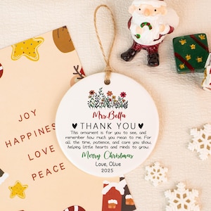 May include: A white ceramic Christmas ornament with a floral design and the text "Mrs. Bella THANK YOU Merry Christmas Love, Olive 2025". The ornament hangs from a twine loop. Other Christmas decorations are in the background.