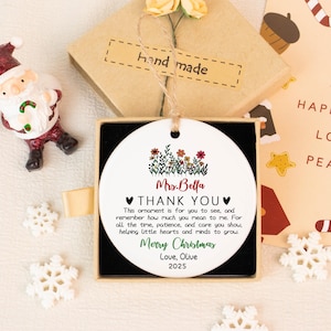 May include: A white ceramic Christmas ornament with floral accents and the text "Mrs. Bella THANK YOU Merry Christmas Love, Olive 2025" in a gift box. The ornament is round and has a string for hanging. A small Santa Claus figurine and snowflakes are also visible.