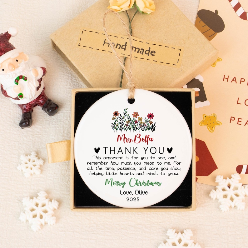 May include: A white ceramic Christmas ornament with floral accents and the text "Mrs. Bella THANK YOU Merry Christmas Love, Olive 2025" in a gift box. The ornament is round and has a string for hanging. A small Santa Claus figurine and snowflakes are also visible.