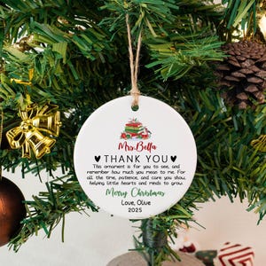 May include: A white ceramic Christmas ornament with a printed message. The ornament has a string for hanging and features text that reads "Mrs. Bella THANK YOU Merry Christmas Love, Olive 2025".