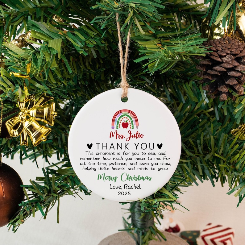May include: A white, round Christmas ornament with a rainbow and apple design. Text reads "Mrs. Julie THANK YOU" and a Christmas message. The ornament hangs from a twine string, displayed on a Christmas tree.