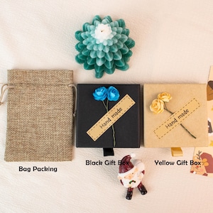 May include: An overhead shot features a teal and white flower-shaped candle, a burlap bag, and two gift boxes. The black box has blue flowers and a "Hand made" label. The yellow box has yellow flowers and a "Hand made" label. A Santa figurine is also present.