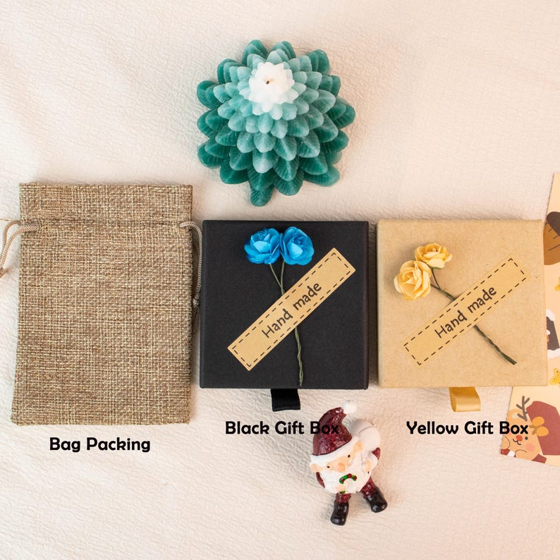 May include: An overhead shot features a teal and white flower-shaped candle, a burlap bag, and two gift boxes. The black box has blue flowers and a "Hand made" label. The yellow box has yellow flowers and a "Hand made" label. A Santa figurine is also present.
