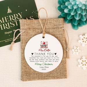 May include: A white ceramic Christmas ornament with a teacher-themed design and the text "Mrs. Bella THANK YOU Merry Christmas Love, Olive 2025". The ornament hangs from a jute string and is presented in a burlap gift bag.