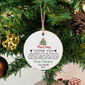 May include: A white, round Christmas ornament with a brown string for hanging. The ornament has text that reads "Miss Daisy THANK YOU" and "Merry Christmas Love, Rachel 2025". The ornament is hanging on a Christmas tree.