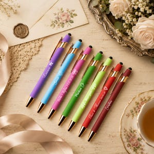 May include: A set of seven retractable ballpoint pens in purple, light blue, magenta, green, lime green, red, and dark red. Each pen features a gold-tone clip, a black stylus tip, and white custom text including names and dates.
