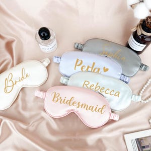 May include: Five satin sleep masks with gold lettering. The masks are in shades of pink, white, light blue, and gray. The text on the masks reads "Bride", "Bridesmaid", "Perla", "Rebecca", and "Sophia".