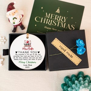 May include: A white ceramic Christmas ornament with a message for a teacher, presented in a black gift box. The ornament has a teacher-themed design and the text "Mrs. Bella THANK YOU". A "Hand made" tag and blue flowers are included.