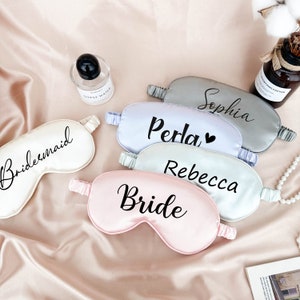 May include: Five satin sleep masks with different names printed on them. The masks are in shades of pink, light blue, and gray. The names on the masks are "Bride", "Rebecca", "Perla", "Sophia", and "Bridesmaid".