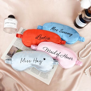 May include: Set of five satin sleep masks with personalized text. The masks are in blue, pink, light blue, and orange. The text on the masks reads "Mrs. Sweeney", "Lydia", "Maid of Honor", "Miss Hay", and a heart symbol.