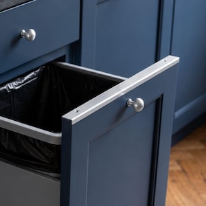 Op de afbeelding: Een uittrekbare keukenlade met een ingebouwde afvalbak voorzien van een zwarte plastic zak. De kast is uitgevoerd in diep marineblauw met een shaker-stijl deurpaneel en een kleine, ronde, zilverkleurige metalen knop als handgreep.