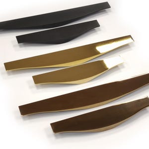May include: A collection of six modern cabinet pulls in various metallic finishes. Two are matte black, two are gold, and two are bronze. Each pull has a curved, elongated shape with a flat, rectangular end.