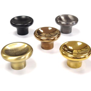 Pode incluir: Cinco puxadores de armário em vários acabamentos metálicos são exibidos em um fundo branco. Os puxadores incluem tons de preto, bronze, latão e ouro. Cada puxador tem um topo arredondado e uma base cilíndrica curta.