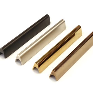 Può includere: Quattro maniglie per mobili in fila, ciascuna con una finitura metallica diversa. Le finiture includono nero opaco, argento spazzolato, oro lucido e bronzo. Ogni maniglia ha una forma a barra rettangolare con un'impugnatura curva e incassata.