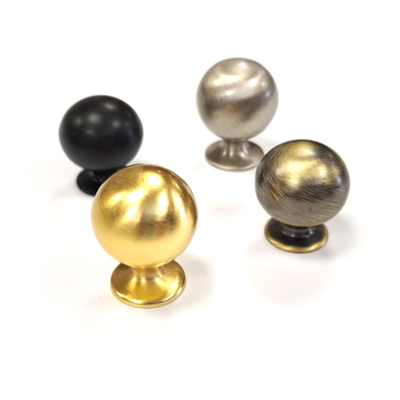 Op de afbeelding: Vier bolvormige kastknoppen in verschillende metallic afwerkingen. De knoppen hebben een kleine, ronde basis. Afwerkingen zijn onder meer zwart, goud, zilver en brons. Geschikt voor meubel- en woondecoratieprojecten. Elke knop heeft een diameter van ongeveer 3 cm.