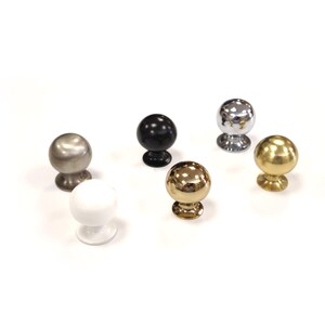 May include: Six spherical cabinet knobs in various finishes: silver, black, chrome, gold, and white. Each knob has a round base. The knobs are arranged on a white surface, showcasing their different colours and textures.