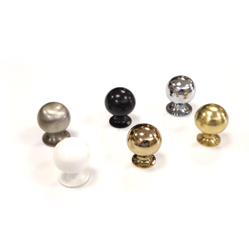 May include: Six spherical cabinet knobs in various finishes: silver, black, chrome, gold, and white. Each knob has a round base. The knobs are arranged on a white surface, showcasing their different colours and textures.