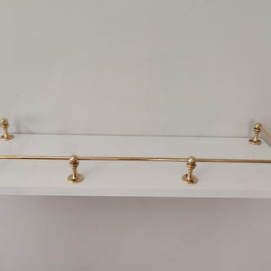 May include: A white floating shelf with brass-colored metal railing. The shelf has a rectangular shape with rounded brass accents at each corner and along the front edge. The shelf is designed for wall mounting.
