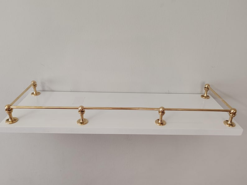 May include: A white floating shelf with brass-colored metal railing. The shelf has a rectangular shape with rounded brass accents at each corner and along the front edge. The shelf is designed for wall mounting.