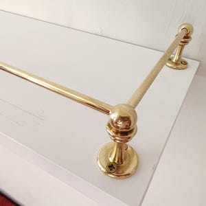 May include: A gold-colored towel bar with a rounded design. The bar is mounted on a white surface and has a classic, elegant appearance. The bar is made of metal and has a polished finish.