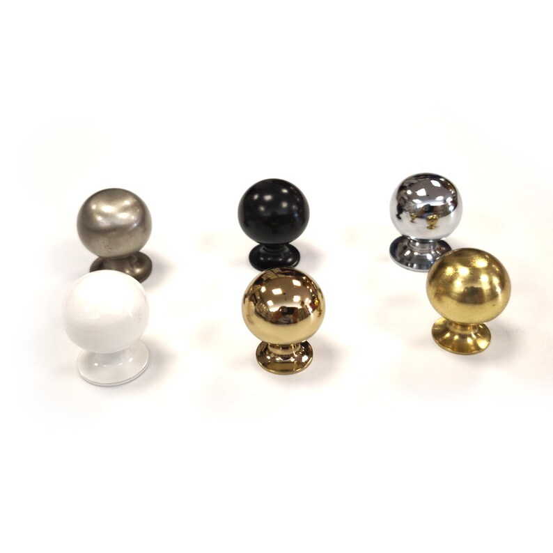 May include: Six spherical cabinet knobs in different finishes are displayed. The knobs include silver, black, gold, chrome, and white. Each knob has a round top and a decorative base. The knobs are shown on a white surface.