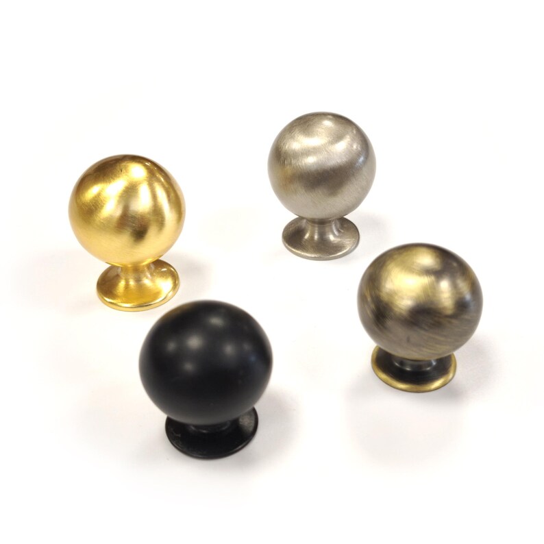 Op de afbeelding: Vier decoratieve kastknoppen in verschillende metallic afwerkingen. De knoppen hebben een bolvormig ontwerp met een kleine, ronde basis. De afwerkingen omvatten goud, zilver, zwart en een bronsachtige kleur.