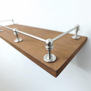 May include: A wooden shelf with a dark brown finish and brushed nickel railing. The railing is composed of horizontal bars and decorative vertical supports. The shelf is mounted on a white wall.