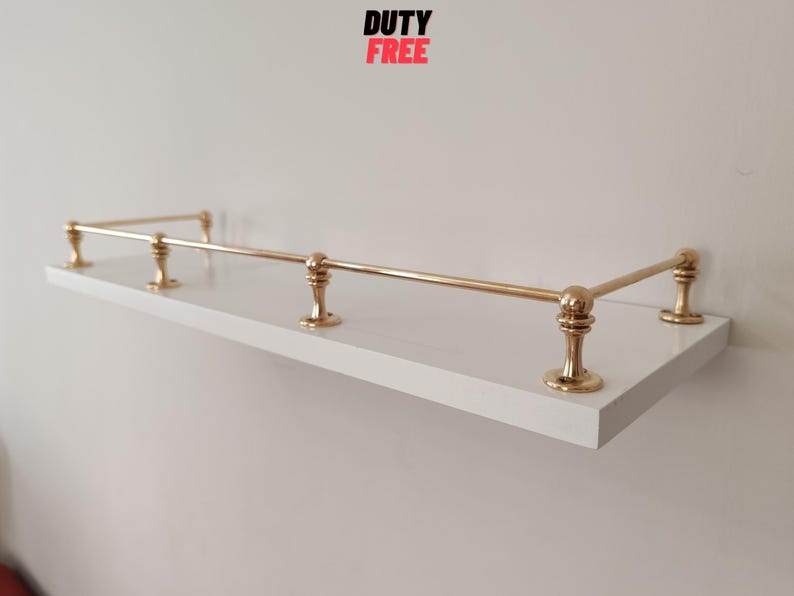 May include: A white floating shelf with gold-colored metal railing and supports. The shelf has a rectangular shape and is mounted on a white wall. The text "DUTY FREE" is in the top center.
